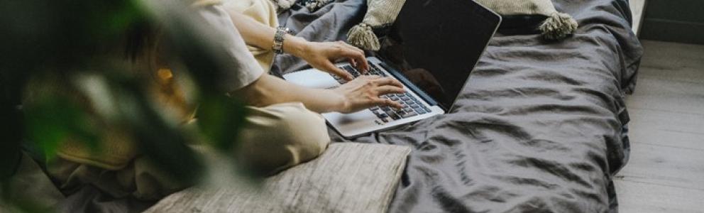Psychologische Onlineberatung: Frau sitzt auf einem Bett und tippt Nachricht in Laptop.