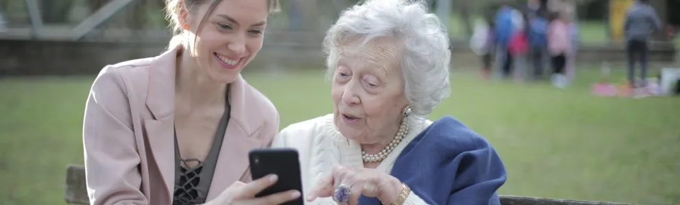 Eine junge und eine alte Frau sitzen auf einer Parkbank und schauen in ein Smartphone. 